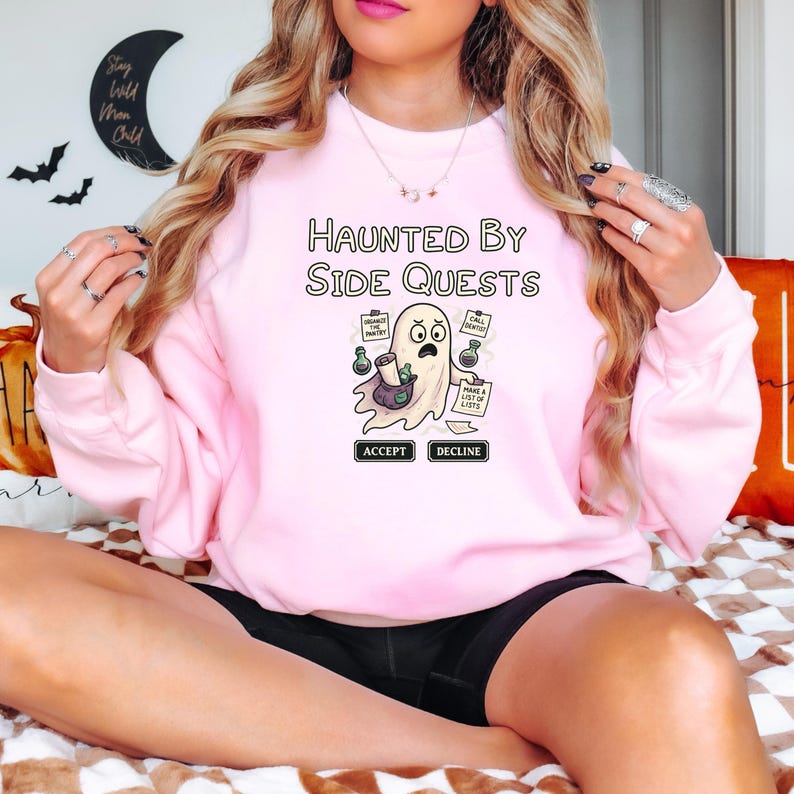May include: A light pink sweatshirt with the text "Haunted By Side Quests" above a cartoon ghost character. The ghost holds a bag and papers with tasks. The word "Accept" and "Decline" are below the ghost.