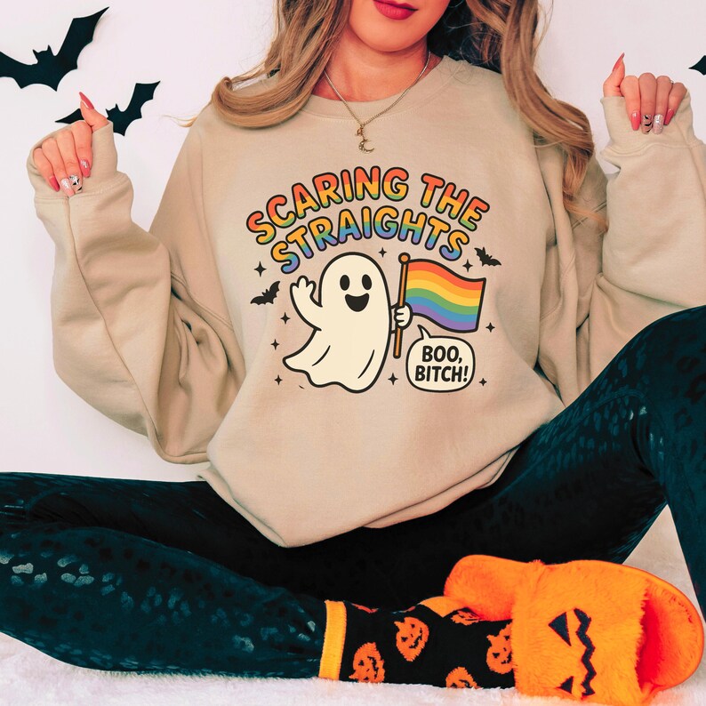 May include: Beige sweatshirt with the text "Scaring the Straights" in rainbow colors, featuring a cartoon ghost holding a rainbow flag. The ghost says "Boo, Bitch!" with bats and stars. The sweatshirt is part of a Halloween-themed collection.