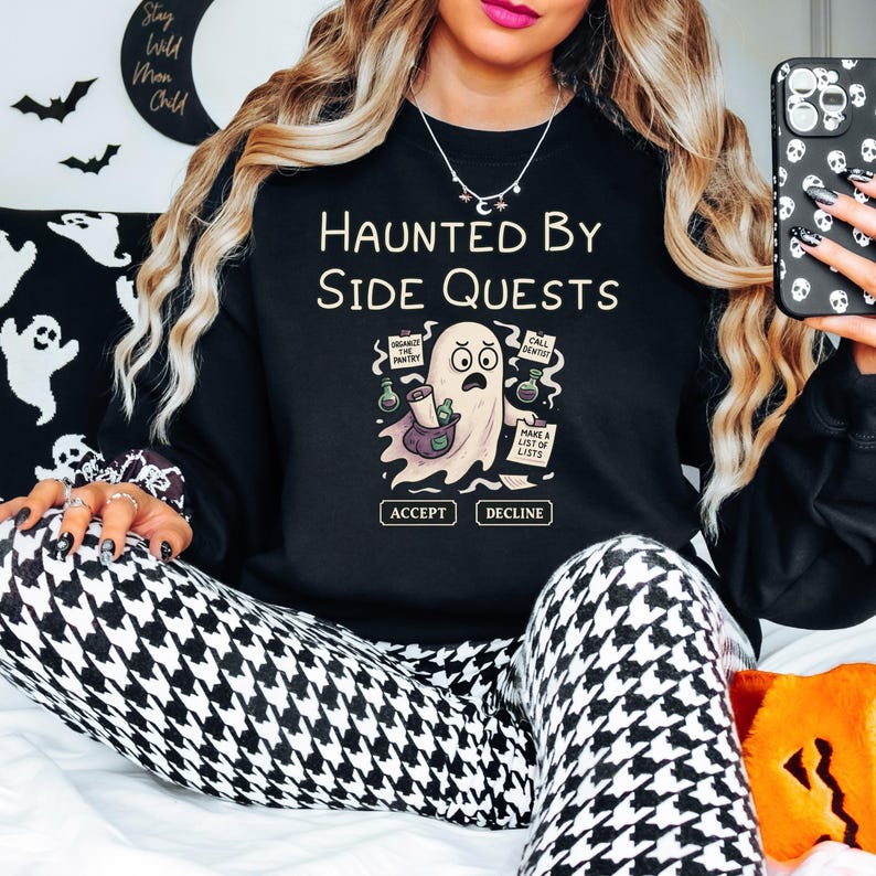 May include: Black sweatshirt with the text "HAUNTED BY SIDE QUESTS" and a cartoon ghost illustration. The ghost is holding a scroll and surrounded by speech bubbles with text. The sweatshirt has the words "ACCEPT" and "DECLINE" below the ghost.