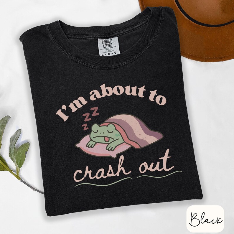 Pu&ograve; includere: T-shirt nero con la scritta "I'm about to crash out" e un'illustrazione di una rana che dorme. Il testo e l'immagine sono in morbide tonalit&agrave; rosa e verde. Un cappello marrone &egrave; visibile sullo sfondo.