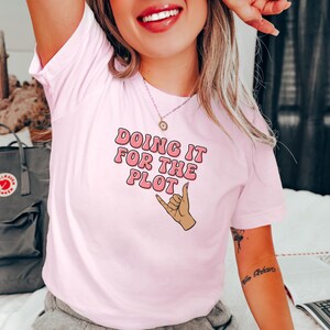 Puede incluir: Camiseta rosa con el texto "Doing it for the plot" en rosa con una mano marrón haciendo un signo shaka.