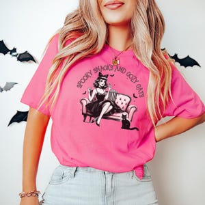 May include: A pink t-shirt with a graphic of a woman sitting on a couch with a cat. The text on the shirt reads "Spooky Snacks and Cozy Cats".