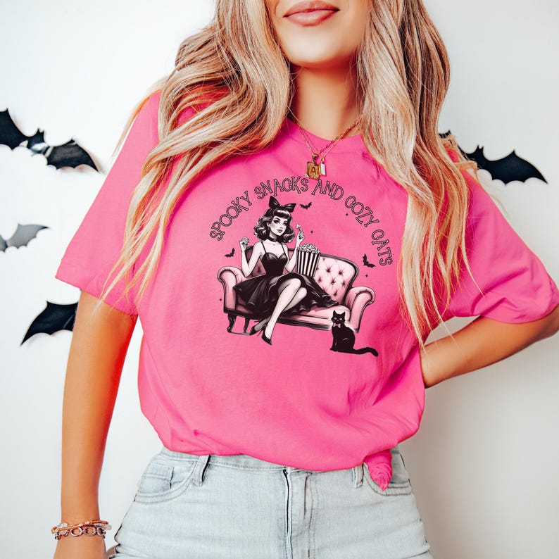May include: A pink t-shirt with a graphic of a woman sitting on a couch with a cat. The text on the shirt reads "Spooky Snacks and Cozy Cats".