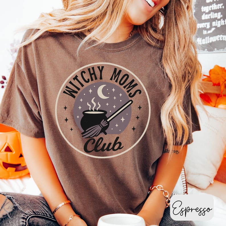 May include: Espresso-colored t-shirt featuring a circular graphic with the text "Witchy Moms Club." The design includes a cauldron, broom, crescent moon, and stars. The shirt is a comfortable, casual style.
