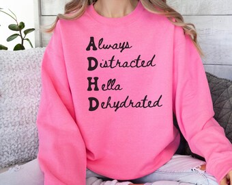 May include: A bright pink sweatshirt with the text "Always Distracted Hella Dehydrated" in black script. The sweatshirt is a crew neck style and appears to be made of a soft material. The overall theme is casual wear.