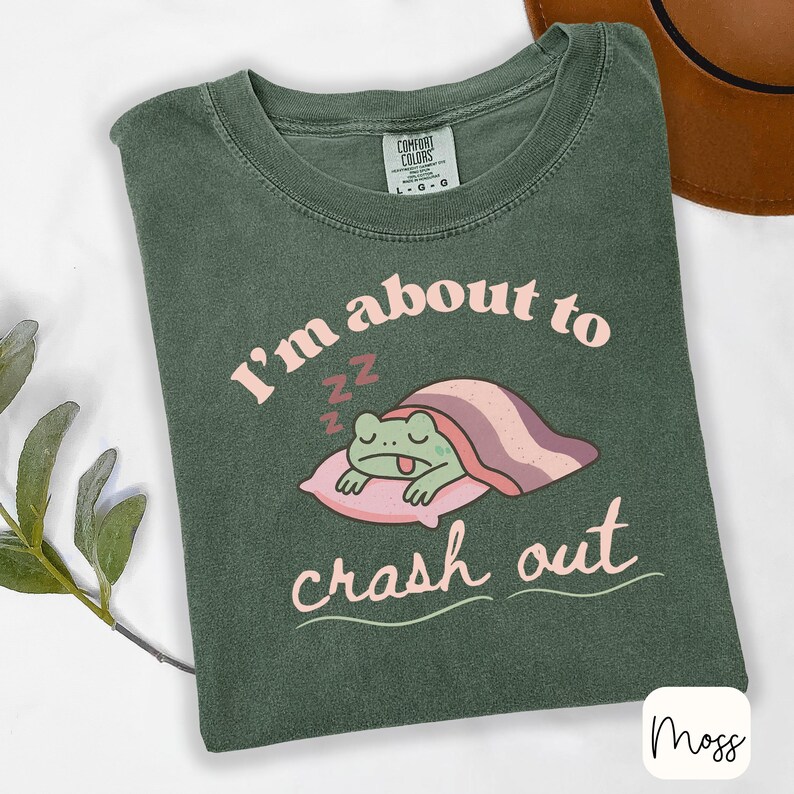 Pu&ograve; includere: T-shirt verde muschio con la scritta "I'm about to crash out" e una grafica di una rana che dorme. La rana &egrave; su un cuscino rosa sotto una coperta a righe. La maglietta ha un girocollo e maniche corte.