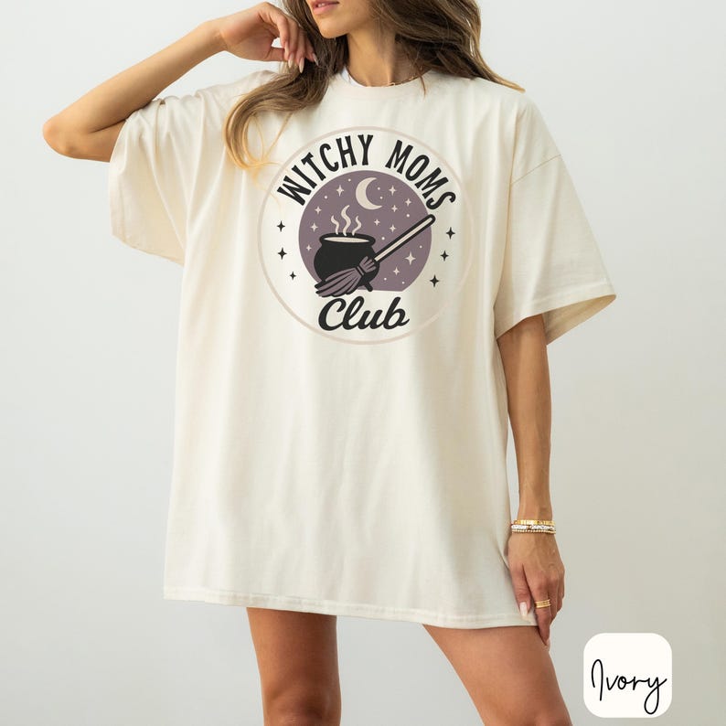 May include: Ivory-colored t-shirt featuring a circular graphic with the text "Witchy Moms Club." The graphic includes a cauldron, broom, moon, and stars. The shirt has short sleeves and a relaxed fit.