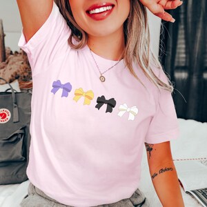 May include: A pink t-shirt with a row of four colorful bows printed on the front. The bows are purple, yellow, black, and white.