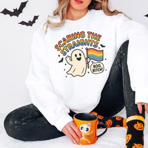 May include: White sweatshirt with the text "Scaring the Straights" and a cartoon ghost holding a rainbow flag. The ghost says "Boo, Bitch!" in a speech bubble. Black leggings and an orange Halloween mug are also visible.