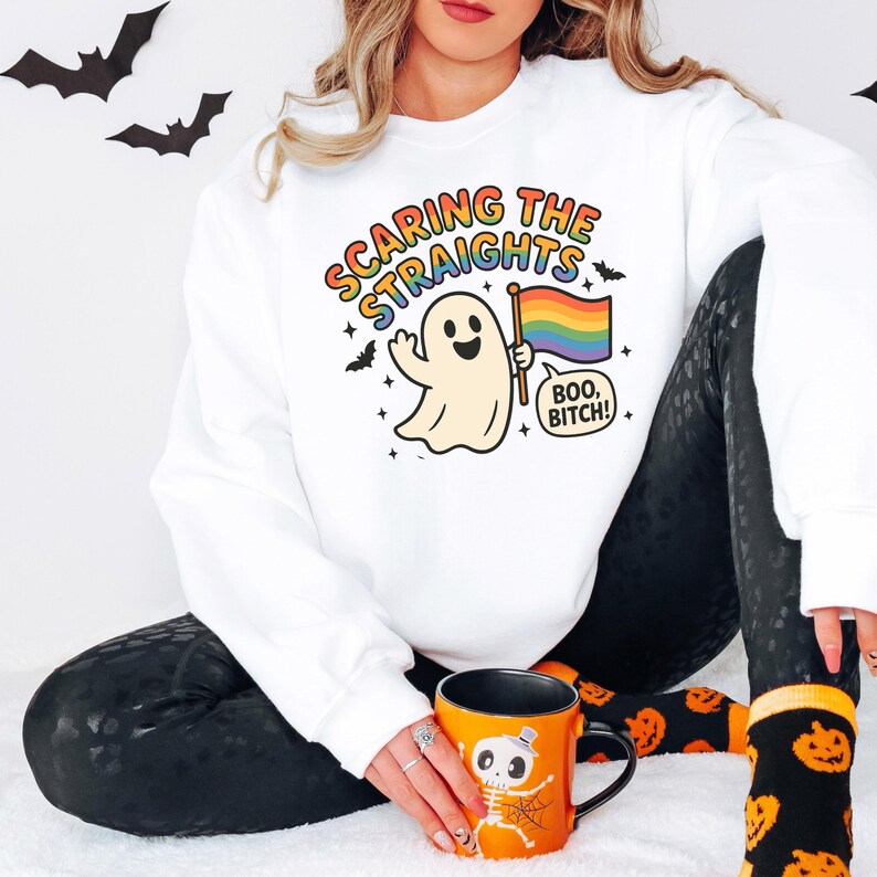May include: White sweatshirt with the text "Scaring the Straights" and a cartoon ghost holding a rainbow flag. The ghost says "Boo, Bitch!" in a speech bubble. Black leggings and an orange Halloween mug are also visible.