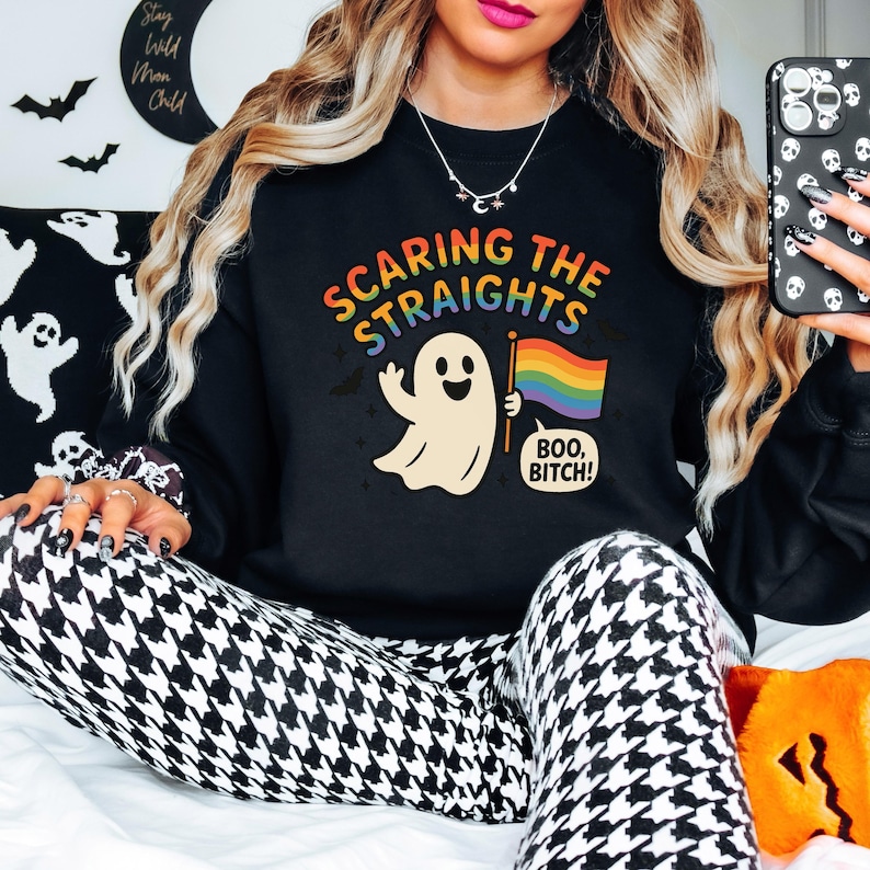 May include: Black sweatshirt with the text "Scaring the Straights" in rainbow colors. A cartoon ghost holds a rainbow flag and the words "Boo, Bitch!" are below. The person is wearing black and white houndstooth pants.