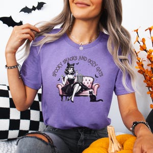 May include: Purple t-shirt with a graphic of a woman in a black dress sitting on a pink couch with a black cat. The text on the shirt reads "Spooky Snacks and Cozy Cats".