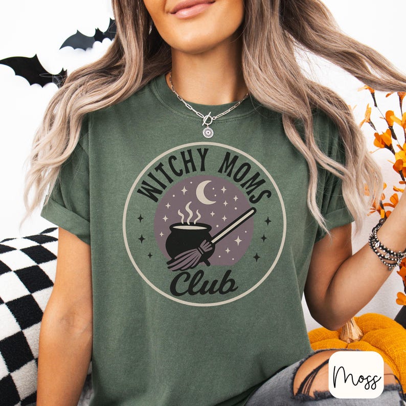 May include: Olive green t-shirt with a circular graphic that reads "Witchy Moms Club." The graphic features a cauldron, broom, moon, and stars. The shirt has rolled-up sleeves and is made of a soft material.