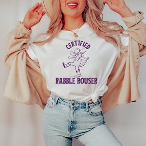 May include: White t-shirt with a purple graphic of a cat wearing a cowboy hat and boots. The text "CERTIFIED RABBLE ROUSER" is printed below the cat.