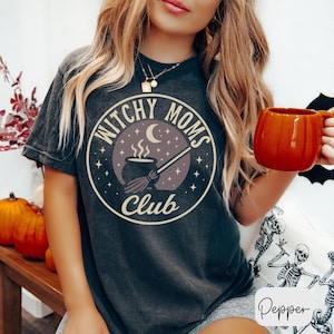 May include: Dark gray t-shirt with a circular graphic that reads "Witchy Moms Club." The design features a cauldron, broom, moon, and stars. The person is holding an orange mug. Pumpkins are visible in the background.