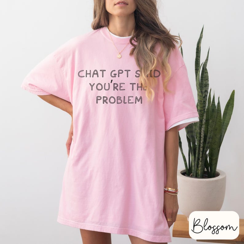 May include: A light pink t-shirt with the text "CHAT GPT SAID YOU'RE THE PROBLEM" in black. The shirt has white trim on the sleeves and neckline. The model is wearing gold jewelry.
