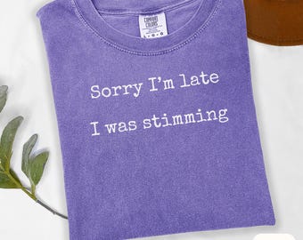 May include: A violet-colored t-shirt with the text "Sorry I'm late I was stimming" in white. The shirt has a crew neck and a tag that says "Comfort Colors". A brown hat is partially visible in the background.