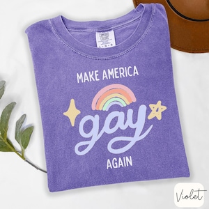 May include: A violet-colored t-shirt with the text "MAKE AMERICA GAY AGAIN" in white, featuring a rainbow and stars graphic. The shirt is made of a soft material and has a crew neck.