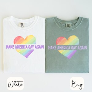 May include: Two t-shirts, one white and one bay green, each featuring a rainbow heart design. The text "MAKE AMERICA GAY AGAIN" is printed across the heart. The shirts are made of a soft material.