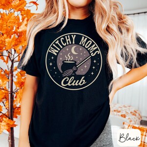 May include: Black t-shirt with a circular graphic that reads "Witchy Moms Club." The design features a cauldron, moon, stars, and a broom. The shirt is a comfortable fit and suitable for casual wear.