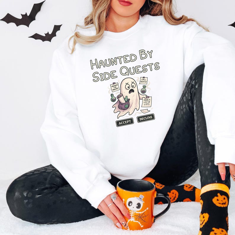 May include: White sweatshirt with the text "HAUNTED BY SIDE QUESTS" and a cartoon ghost graphic. The ghost holds potions and has speech bubbles that say "ACCEPT" and "DECLINE". An orange Halloween mug with a skeleton design is also visible.