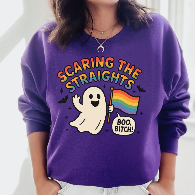 May include: A purple sweatshirt with the text "SCARING THE STRAIGHTS" in rainbow colors. A cartoon ghost holds a rainbow flag and a speech bubble that says "BOO, BITCH!". The design includes bats and stars.