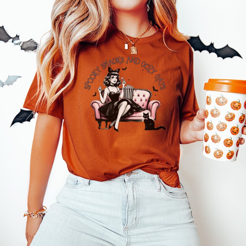 May include: A woman wearing a rust-colored t-shirt with the text "Spooky Snacks and Cozy Cats" and a black and white illustration of a woman sitting on a pink couch with a black cat. She is holding a bucket of popcorn.