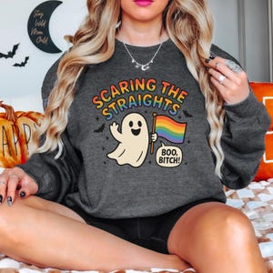 May include: Dark gray sweatshirt with the text "SCARING THE STRAIGHTS" in rainbow colors. A cartoon ghost holds a rainbow flag and a speech bubble says "BOO, BITCH!" Bats and stars are also featured.