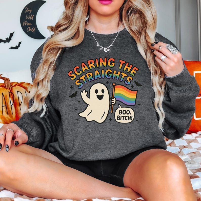 May include: Dark gray sweatshirt with the text "SCARING THE STRAIGHTS" in rainbow colors. A cartoon ghost holds a rainbow flag and a speech bubble says "BOO, BITCH!" Bats and stars are also featured.