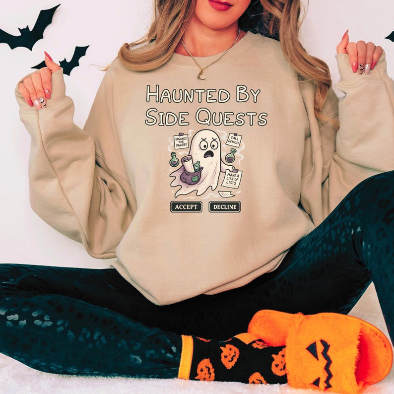 May include: Beige sweatshirt with the text "Haunted By Side Quests" and a cartoon ghost graphic. The ghost holds a bag and lists, with "Accept" and "Decline" buttons below. Black leggings and pumpkin-themed socks are also visible.