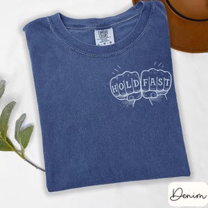 May include: A denim blue Comfort Colors t-shirt with the words "HOLD FAST" in white, stylized as two fists. The shirt is folded and has a tag at the neckline. A brown hat is partially visible in the background.