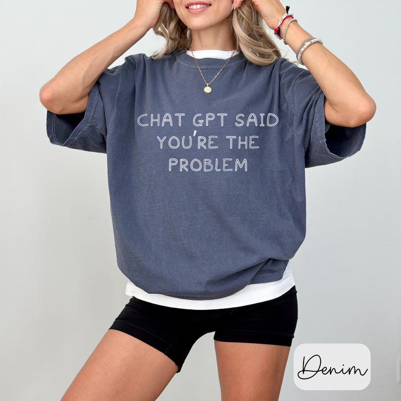 May include: A denim blue t-shirt with the text "CHAT GPT SAID YOU'RE THE PROBLEM" in white. The shirt has short sleeves and a relaxed fit. The model is wearing black shorts.