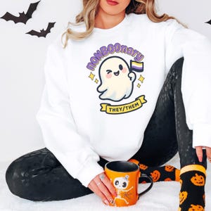 May include: White sweatshirt with a cartoon ghost holding a non-binary pride flag. The ghost has a winking face and is surrounded by stars. The text "NONBOONARY" and "THEY/THEM" are also featured on the sweatshirt. A Halloween-themed mug is also visible.