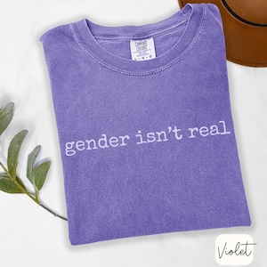 May include: A violet-colored t-shirt with the text "gender isn't real" in white. The shirt is folded and has a Comfort Colors tag. A brown hat is visible in the background.