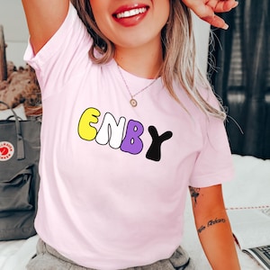 May include: A pink t-shirt with the word "ENBY" written in colorful bubble letters. The letters are yellow, white, purple, and black.