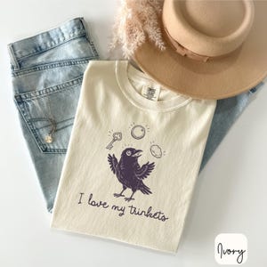 May include: An ivory t-shirt featuring a dark purple graphic of a crow surrounded by a key, a coin, and a stone, with the text "I love my trinkets" printed below. The shirt is layered with light blue denim jeans and a tan wide-brimmed hat.