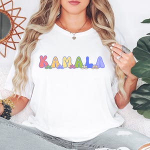 May include: White t-shirt with the word "KAMALA" in rainbow colored letters with flowers underneath.