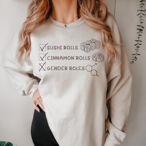 May include: Beige sweatshirt with the text "Sushi Rolls," "Cinnamon Rolls," and "Gender Roles." The sushi rolls and cinnamon rolls are checked, while gender roles are crossed out. The design includes illustrations of sushi rolls and a cinnamon roll.