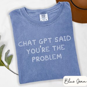 May include: A blue jean colored t-shirt with the text "CHAT GPT SAID YOU'RE THE PROBLEM" in white. The shirt has a crew neck and a Comfort Colors tag. A brown hat is partially visible in the background.