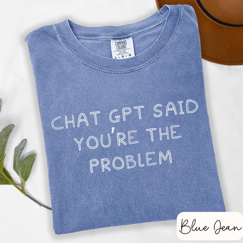 May include: A blue jean colored t-shirt with the text "CHAT GPT SAID YOU'RE THE PROBLEM" in white. The shirt has a crew neck and a Comfort Colors tag. A brown hat is partially visible in the background.
