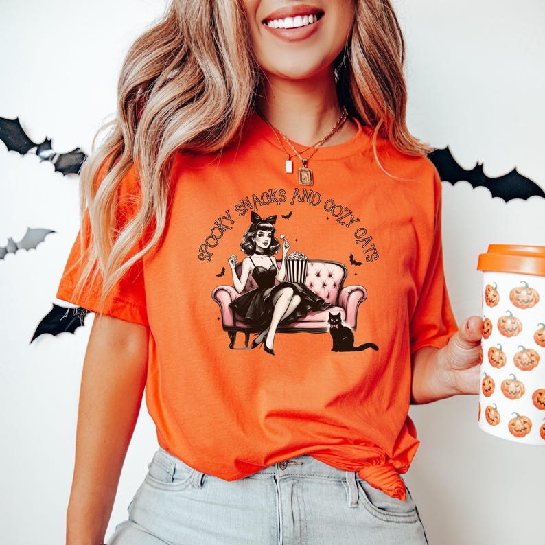 May include: Orange t-shirt with a graphic of a woman sitting on a couch with a cat and popcorn. The text on the shirt reads "Spooky Snacks and Cozy Cats".