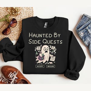 May include: Black crewneck sweatshirt with the text "Haunted By Side Quests" and a cartoon ghost illustration. The ghost holds a potion bottle and a list, with options to "Accept" or "Decline" side quests. A fun, spooky design.