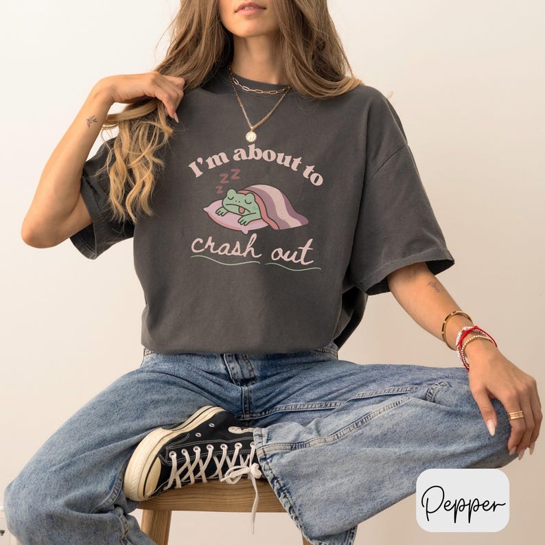 Pu&ograve; includere: T-shirt grigio scuro con la scritta "I'm about to crash out" e un ranocchio dei cartoni animati che dorme su un cuscino. La rana &egrave; verde, il cuscino &egrave; rosa e la coperta &egrave; a righe rosa e viola.