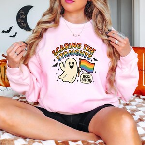 May include: A light pink sweatshirt with the text "Scaring the Straights" above a cartoon ghost holding a rainbow flag. The ghost has a smiling face and is surrounded by bats and stars. The phrase "Boo, Bitch!" is below the ghost.