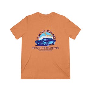 May include: Orange t-shirt with a graphic design featuring a blue classic car, a sun, and the text "TAKE THE HIGHWAY THROUGH THE GREAT DIVIDE". The shirt has short sleeves and a crew neck.