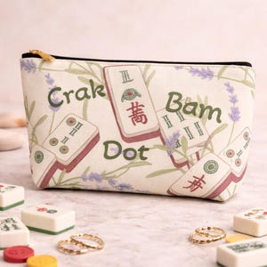 Op de afbeelding: Crèmekleurige etui met ritssluiting met een Mahjong-tegelpatroon en de woorden "Crak", "Bam" en "Dot". De etui heeft een zwarte ritssluiting en een goudkleurige ritstrekker. Lavendeltakken en Mahjong-tegels zijn op de etui gedrukt.