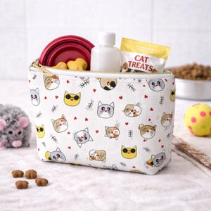 May include: A white canvas zippered pouch with a cat face pattern, holding a red collapsible bowl, treats, and a bottle. A yellow ball and a plush mouse toy are nearby. The pouch is designed for cat supplies.