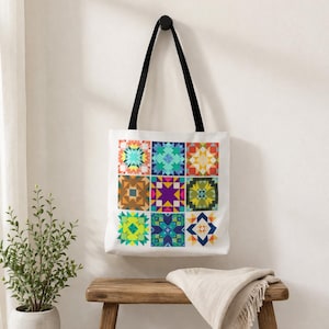 May include: White tote bag with a black handle, featuring nine colorful geometric quilt block designs. The blocks showcase various patterns in shades of blue, green, orange, yellow, and purple. The bag hangs on a black hook against a white wall.
