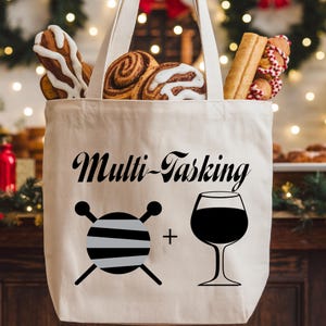 May include: A cream-colored tote bag filled with baked goods. The bag features the text "Multi-Tasking" with a graphic of yarn and a wine glass. The bag has cream-colored handles.