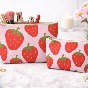 May include: Two pink cosmetic bags with a repeating pattern of red strawberries with green leaves. The larger bag is filled with makeup brushes and other beauty products. Both bags have gold zippers.